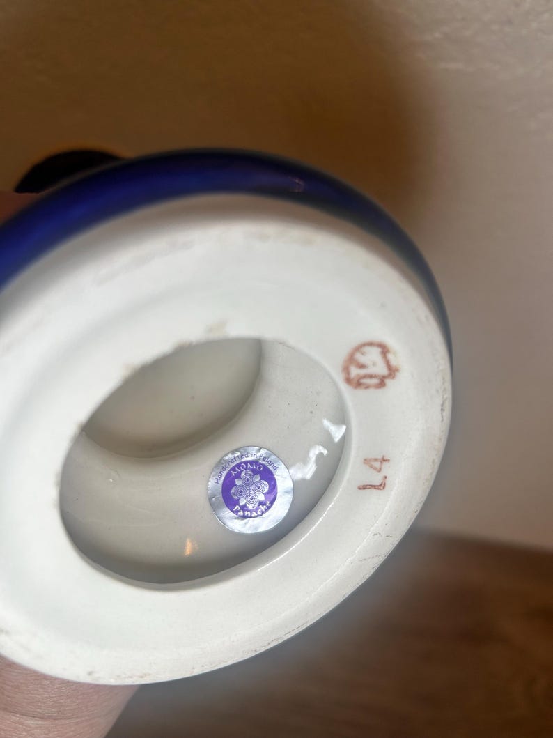 May include: A ceramic vase with a white base and a dark blue rim. The bottom of the vase has a sticker with text and a number 14 in brown ink. The vase is handcrafted.