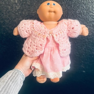 May include: A vintage doll with a peach-colored face and blue eyes, wearing a pink dress and a crocheted pink sweater. The dress has a white lace trim. The doll is being held up against a dark background.