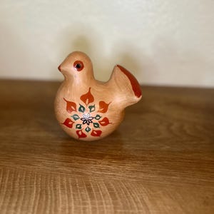 May include: A ceramic bird figurine in shades of tan, red, and green. The bird has a rounded body with a small tail and a detailed floral design on its front. The bird's eye is a small red dot.