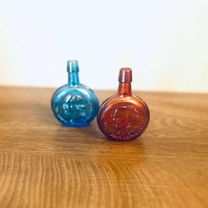 May include: Two antique glass bottles, one blue and one red-orange, with embossed portraits and text. The bottles have a round body and a narrow neck. The bottles are displayed on a wooden surface.