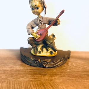 May include: A figurine of a seated character wearing a grey cap and long-sleeved shirt, playing a reddish-brown stringed instrument. The character has large eyes and is positioned on a dark brown, curved base with decorative carved scrollwork details.