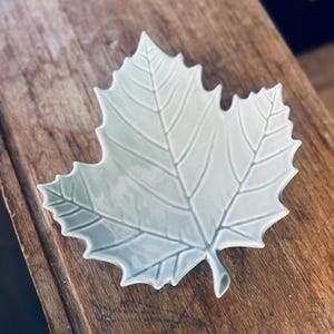 May include: A light green ceramic leaf-shaped dish. The dish has detailed veining and a slightly textured surface. The leaf dish is a decorative item, suitable for serving small items or as a decorative accent piece. The dish is on a wooden surface.