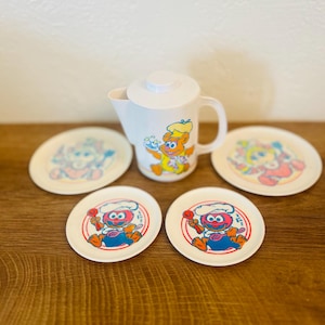 Puede incluir: Una jarra de plástico blanco con tapa y asa, con un oso de dibujos animados como chef. Cuatro platos pequeños con personajes de dibujos animados rodean la jarra. Los platos y la jarra están decorados con ilustraciones coloridas.