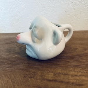 May include: A white ceramic creamer shaped like a human face with a prominent nose, a small pink-painted tip on the nose, closed eyes, and a defined mouth. The vessel features a curved handle on the side and a smooth, glossy finish throughout.