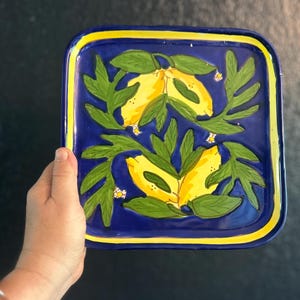 May include: Square ceramic serving tray with a vibrant design. The tray features a deep blue background with a yellow border. The center showcases a lemon and leaf motif in yellow and green, adding a touch of nature-inspired decor.