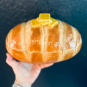 May include: A ceramic bread dish with a glossy, glazed finish. The dish is shaped like a loaf of bread, with vertical lines and a golden-brown color. It is topped with several yellow butter pats.