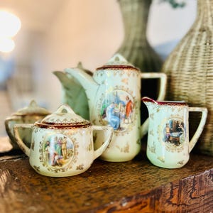 May include: A vintage porcelain tea set with a cream-colored base, featuring detailed illustrations and gold accents. The set includes a teapot, creamer, sugar bowl, and a small pitcher, all with a classic design. The tea set is displayed on a wooden surface.
