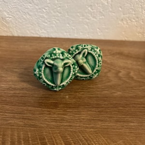 May include: Two green ceramic sheep-shaped salt and pepper shakers. Each shaker features a sheep head with a detailed face and a decorative border. The shakers are on a wooden surface.