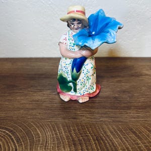 May include: A ceramic figurine of a woman wearing a straw hat and glasses, holding a large blue flower. The woman is dressed in a white dress with a floral pattern. The figurine is on a wooden surface.