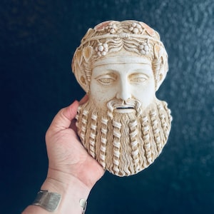 May include: A cream-colored, sculpted bust of a bearded man with a laurel wreath. The bust has detailed facial features and a textured beard. The sculpture is held against a dark blue background.