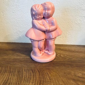 Könnte beinhalten: Eine Vintage-Figur aus rosa Keramik, die zwei sich umarmende Figuren darstellt. Die Figuren sind stilisiert mit abgerundeten Merkmalen und tragen passende Outfits. Die Figur steht auf einem kreisförmigen Sockel und hat ein glänzendes Finish. Das Gesamtthema ist romantisch und sentimental.