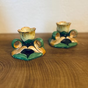 Pode incluir: Dois castiçais de cerâmica com um design floral. Os castiçais têm um esquema de cores amarelo e verde, com um topo em forma de flor e detalhes decorativos. Os castiçais são colocados numa superfície de madeira.