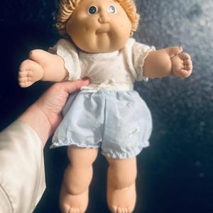 May include: A vintage doll with a round face, blonde yarn hair, and green eyes. The doll is wearing a white top with lace trim and light blue shorts. The doll is being held by a person.
