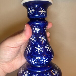 May include: A blue ceramic candlestick with a white top and snowflake pattern. The candlestick has a bulbous base and a slender, elongated body. The snowflake design is white and evenly spaced across the blue surface. The top is white.