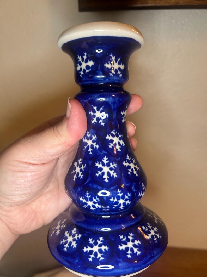 May include: A blue ceramic candlestick with a white top and snowflake pattern. The candlestick has a bulbous base and a slender, elongated body. The snowflake design is white and evenly spaced across the blue surface. The top is white.