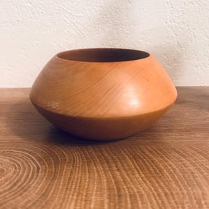 May include: A handcrafted wooden bowl with a rounded, asymmetrical shape. The bowl is made of light brown wood with visible grain patterns. The interior is a darker shade of brown. The bowl sits on a wooden surface.