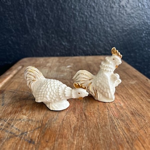 May include: Two ceramic rooster figurines. The roosters are white with gold accents on their combs, beaks, and tail feathers. One rooster is standing upright, while the other is in a downward position. The figurines are on a wooden surface.