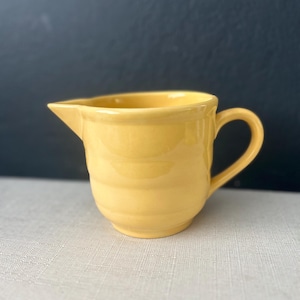 May include: A small, yellow ceramic creamer with a handle and a spout. The creamer has a rounded body with horizontal ridges and sits on a light-colored surface. The background is a dark, solid color.