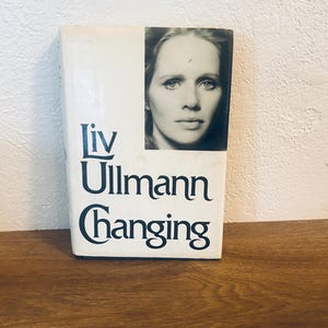 以下が含まれることがあります： リヴ・ウルマン著「Changing」というタイトルのハードカバー本。本の表紙は白で、黒い文字と女性の白黒ポートレートが描かれています。本は木製の表面に置かれています。