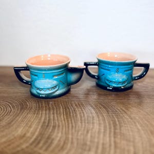 Pode incluir: Duas canecas de cerâmica com um design de tubarão. As canecas são turquesas com alças pretas e uma cara de tubarão na frente. O interior das canecas é cor de pêssego. As canecas estão sobre uma superfície de madeira.