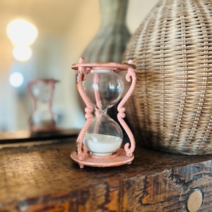 Op de afbeelding: Een kleine, roze zandloper met heldere glazen bollen en wit zand. De zandloper heeft een decoratief, touwachtig ontwerp en staat op een houten oppervlak. Een geweven mand en vaas staan op de achtergrond.