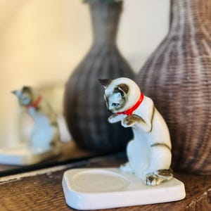 Puede incluir: Figura de gato siamés de cerámica con collar rojo, sobre una base cuadrada blanca. El gato está en una pose juguetona, con las patas delanteras levantadas. La figura está sobre una superficie de madera.