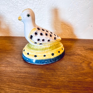 Könnte beinhalten: Keramikvogel-Figur mit weißem Körper, gelbem Schnabel und schwarz-gelb gemusterten Flügeln. Der Vogel sitzt auf einem Sockel mit blauen, gelben und grünen Streifen und schwarzen Punkten. Dekorativer Artikel.