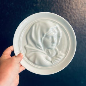 Könnte beinhalten: Dekorative weiße Keramikplatte mit einem Flachrelief einer Frau mit drapierter Kopfbedeckung. Die Platte hat einen erhabenen Rand und eine glatte, glänzende Oberfläche. Das Design ist auf der Platte zentriert.
