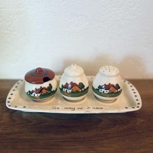 May include: A ceramic condiment set featuring a lidded jar, a shaker, and a container with a lid, each adorned with a house motif. The set rests on a rectangular tray with the text "Go aisy wi' it now".
