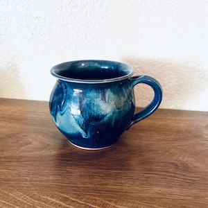 May include: A rounded ceramic mug featuring a glossy glaze with swirling shades of deep navy, royal blue, and lighter turquoise. The mug has a wide, curved body, a flared rim, and a sturdy, matching blue handle attached to the side.