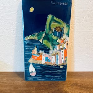 Puede incluir: Azulejo cerámico rectangular que representa la ciudad costera italiana de Portovenere. La obra presenta un cielo azul oscuro con una luna amarilla, una montaña verde y edificios coloridos junto al agua. Un velero está en el agua.