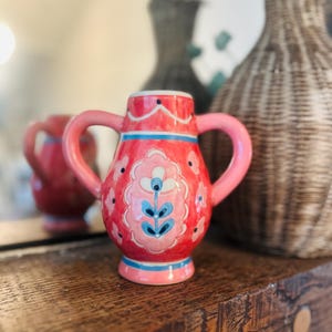 May include: A small, pink ceramic pitcher with two handles. The pitcher is decorated with a floral design in shades of pink, blue, and white. It sits on a wooden shelf, with a blurred background featuring a mirror and decorative vases.