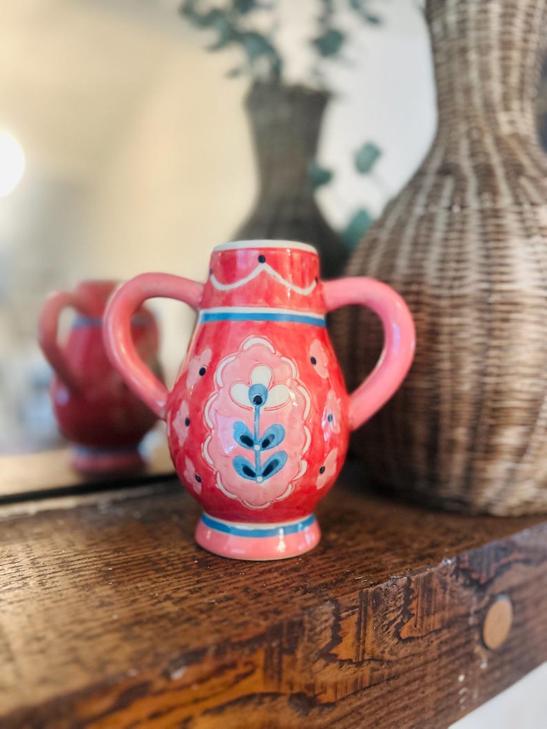 May include: A small, pink ceramic pitcher with two handles. The pitcher is decorated with a floral design in shades of pink, blue, and white. It sits on a wooden shelf, with a blurred background featuring a mirror and decorative vases.