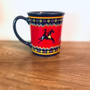 Peut inclure: Mug en céramique rouge avec une anse et un bord noirs. Le mug présente un motif amérindien avec un cavalier à cheval en noir et jaune, sur fond rouge. Le motif comprend des motifs géométriques jaunes, noirs et bleus.