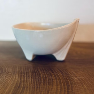 May include: A small, light-colored ceramic bowl with a smooth, glossy finish. The bowl features a unique asymmetrical design with one side extending upward into a pointed, fan-like shape, supported by three short, sturdy feet at the base.