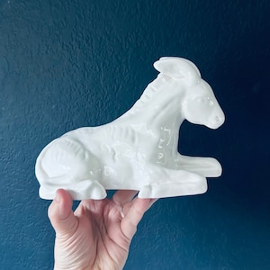 May include: A white ceramic donkey figurine, lying down with its head turned to the side. The donkey has a detailed sculpted design and is a traditional Christmas nativity scene figure.
