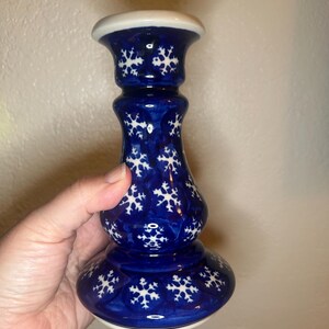 May include: A blue ceramic candlestick with a white snowflake pattern. The candlestick has a wide base, a slender body, and a flared top. The top and base are trimmed with white. The candlestick is held by a hand.