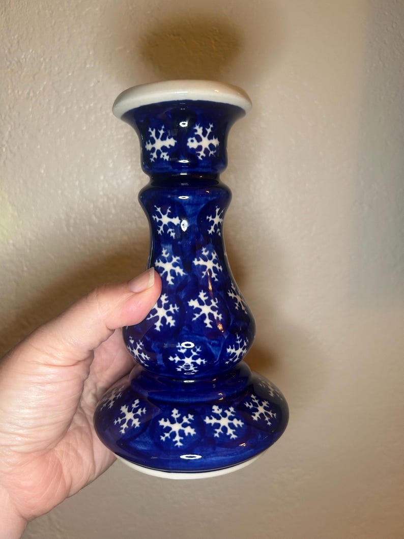 May include: A blue ceramic candlestick with a white snowflake pattern. The candlestick has a wide base, a slender body, and a flared top. The top and base are trimmed with white. The candlestick is held by a hand.