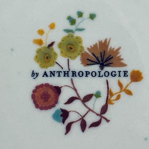 Op de afbeelding: Close-up van een wit oppervlak met een bloemenontwerp en de tekst "by ANTHROPOLOGIE". Het ontwerp bevat geborduurde bloemen in tinten geel, oranje, bruin en blauw, met groene bladeren en stelen. De tekst is donkerblauw.