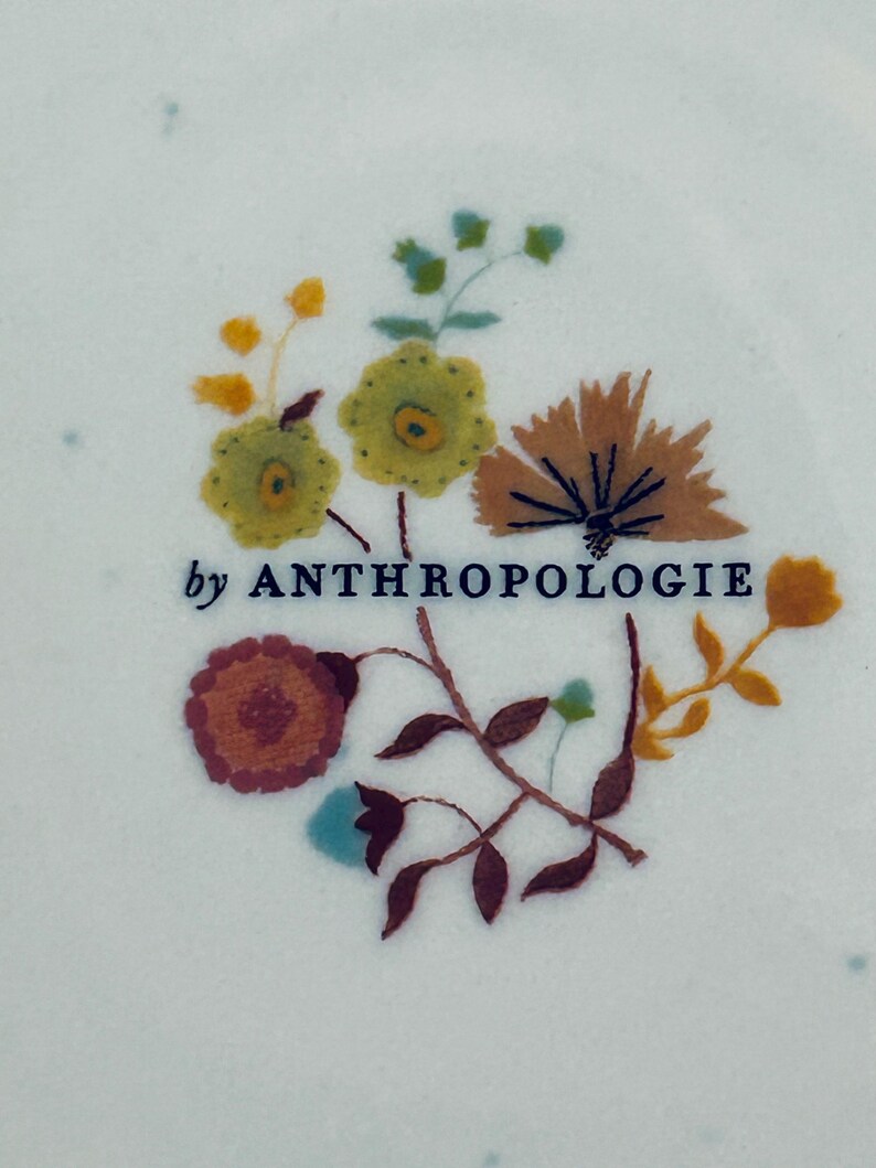 Op de afbeelding: Close-up van een wit oppervlak met een bloemenontwerp en de tekst "by ANTHROPOLOGIE". Het ontwerp bevat geborduurde bloemen in tinten geel, oranje, bruin en blauw, met groene bladeren en stelen. De tekst is donkerblauw.