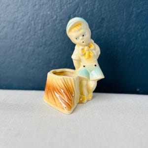 Puede incluir: Un macetero de cerámica con forma de tronco de árbol con una pequeña figura de niño de pie encima. El niño lleva un sombrero azul y blanco, una camisa amarilla, pantalones cortos azules y una corbata de lazo amarilla. El macetero está esmaltado en marrón, amarillo y verde.