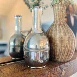 May include: A silver decorative vase with a rounded base and a slender neck, reflecting the surroundings. A similar vase is seen in the background, alongside a woven wicker vase. The vases are displayed on a wooden surface.