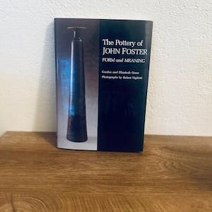 May include: A book titled "The Pottery of John Foster: Form and Meaning" stands on a wooden surface. The cover features a dark blue background with a black and blue pottery vase illustration. The book's title and author's names are in white text.