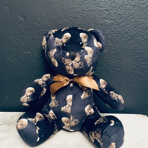 Könnte beinhalten: Ein Plüschteddybär mit dunkelblauem Hintergrund, bedeckt mit einem sich wiederholenden Muster aus Totenköpfen. Der Bär hat eine goldfarbene Schleife um den Hals. Der Bär sitzt auf einer weißen Oberfläche.