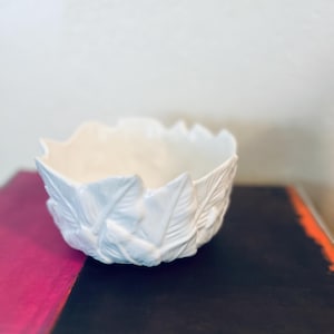May include: A white ceramic bowl with a leaf design. The bowl has a scalloped edge and is resting on a book with a pink and black cover. The bowl's exterior is adorned with detailed, raised leaf patterns.
