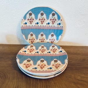 Op de afbeelding: Een set van drie keramische mokken met een gespikkelde, cr&egrave;mekleurige glazuur. Elke mok heeft een ander handgeschilderd bloemmotief in tinten blauw, groen en geel. De mokken hebben een eenvoudige, moderne vorm.