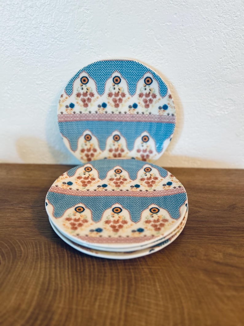 Op de afbeelding: Een set van drie keramische mokken met een gespikkelde, cr&egrave;mekleurige glazuur. Elke mok heeft een ander handgeschilderd bloemmotief in tinten blauw, groen en geel. De mokken hebben een eenvoudige, moderne vorm.