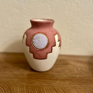 Peut inclure: Un petit vase décoratif avec un corps arrondi et un col étroit. Le vase présente un design bicolore, avec une base blanche et une section supérieure rose. Des formes géométriques dorées et un cercle violet clair avec une bordure dorée ornent la section rose.