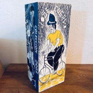 May include: A tall, rectangular box with a blue spine featuring the text "COLECCION 2003." The box's front panel displays a cartoon illustration of a man riding a bicycle, wearing a yellow shirt and a blue hat. The background is black and white.
