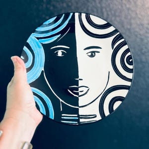 May include: A decorative ceramic plate featuring a stylized face design. The plate is divided into black and white halves, with blue accents. The face has simple lines and circular patterns. The plate is held by a hand.